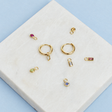 Gold jewelry charms and small components arranged on a white marble tile against a light blue background. The set includes two gold hoop earrings with rectangular clear stone pendants, several gold bezel and teardrop charms in pink, green, blue, and clear stones, and small gold connector rings.
