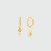 A pair of gold huggie hoop earrings with a small dangling Citrine, shown on a light gray background with a soft reflection.