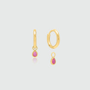 A pair of gold huggie hoop earrings with a small teardrop-shaped Ruby gemstone dangle, shown on a light gray background.