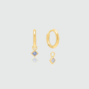 A pair of gold huggie hoop earrings with small diamond-shaped blue gemstone drops, displayed on a light gray background with subtle reflections.