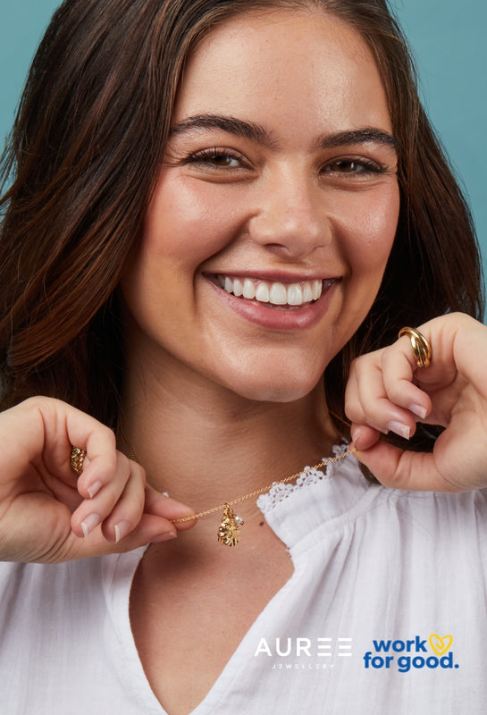 Woman wearing gold jewelry with a blue background, featuring Auree Jewellery and Work For Good Charity branding.