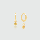 A pair of gold huggie hoop earrings with a small dangling Citrine, shown on a light gray background with a soft reflection.