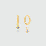 A pair of gold huggie hoop earrings with small diamond-shaped blue gemstone drops, displayed on a light gray background with subtle reflections.
